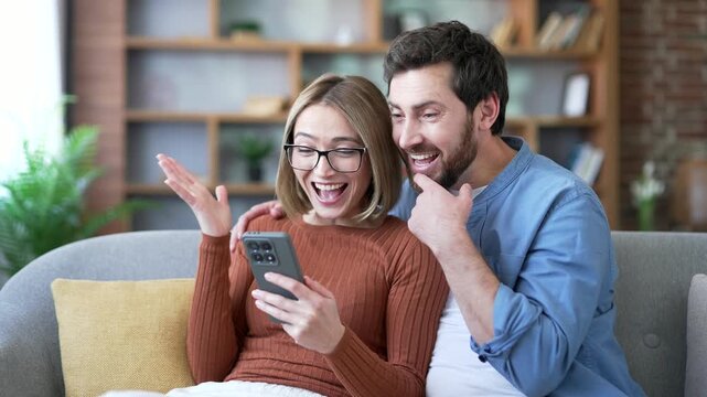 Happy married couple celebrating success reading great news on mobile phone sitting on sofa in living room at home. Smiling joyful husband and wife satisfied with positive good message on smartphone