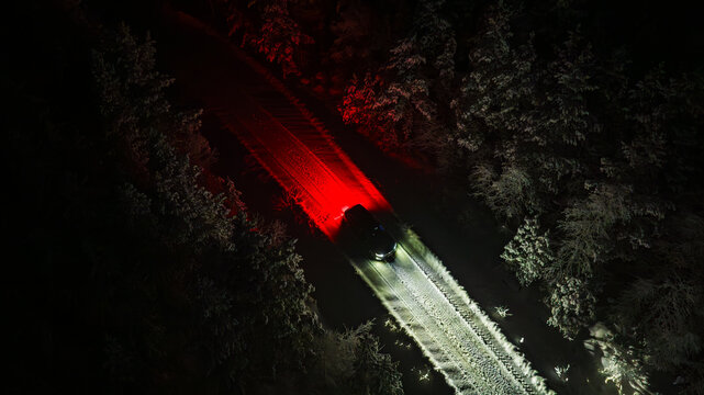 Aerial view of a vehicle with vibrant red and white lights cutting through the dark, snow-dusted forest road, Salla, Lapland, Finland.