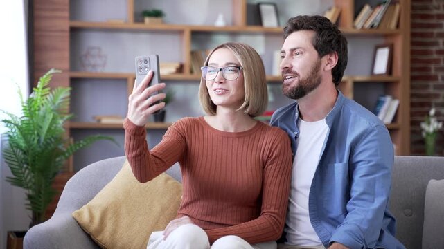 Happy married couple talking on video call using phone while sitting on sofa in living room at home. Cheerful wife and husband have an online meeting with friends, waving hands, joking, having fun, sm