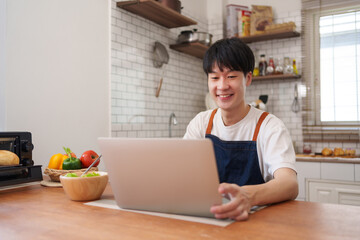 Young man smiling and cooking food at home, watching an online recipe tutorial on laptop in the kitchen