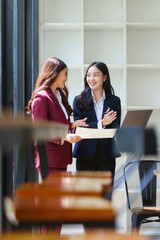Two smiling Asian businesswomen having a professional discussion, reviewing documents and a laptop in a modern office environment