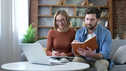 Family couple watching video call training notes in notebook looking at laptop computer screen sitting on sofa in living room at home. Husband and wife listening remote training, webinar or course