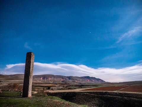 View of standing stone monolith against a backdrop of rolling hills and distant windmills under a vast sky, Goldendale, Washington, United States.