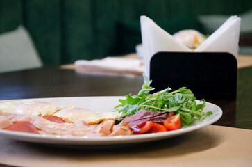 Plate of fried eggs served with sliced bacon, fresh greens, and tomatoes on a white plate.
