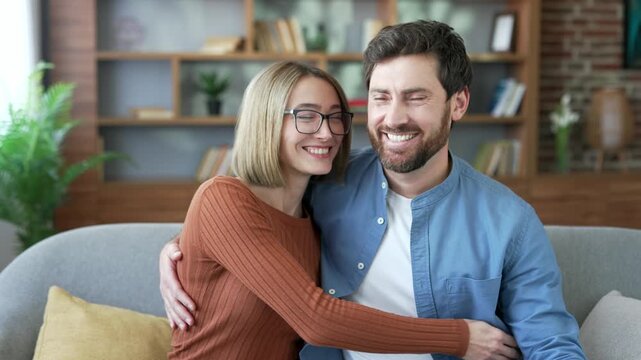 Portrait of happy married couple hugging sitting on sofa in living room posing looking at camera. Loving wife and husband hug and smile. Cheerful man and woman together at home. Head shot of family