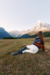 Fototapeta premium Woman in a meadow relaxing and watching mountains and dramatic landscape, nature escape with sweater and boots, outdoor travel scene in a peaceful alpine valley panorama.