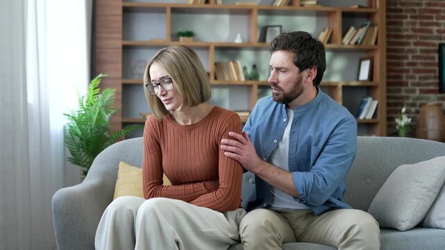 Unhealthy woman feels a sharp pain in her stomach while sitting on sofa in living room at home. Compassionate husband supports, comforts and reassures his wife. Unwell female has menstruation period