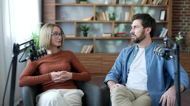 Confident man and woman talking using microphones while recording a podcast sitting in modern studio. Two bloggers looking at each other discuss, have a conversation, interview for video blog content