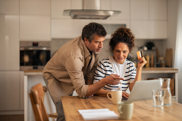 Couple Shopping Online At Kitchen Table With Laptop And Credit Card