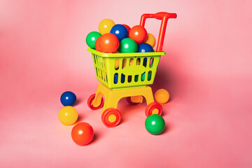 A small shopping cart is filled with colorful balls. The cart sits on a flat surface while more balls are scattered nearby. The backdrop is bright pink.