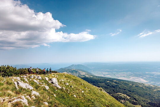 View of hikers silhouetted on a grassy peak against a stunning vista of distant mountains and hazy valleys under a vast, bright sky, Mala Ucka, Primorje-Gorski Kotar County, Croatia.