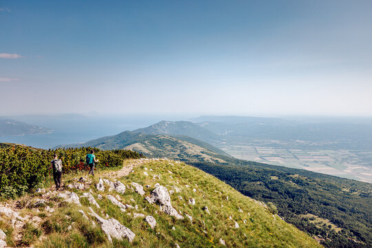View of hikers traversing the vibrant green, rocky terrain of a mountain ridge under a vast blue sky, with distant hazy hills and sea, Mala Ucka, Primorje-Gorski Kotar County, Croatia.