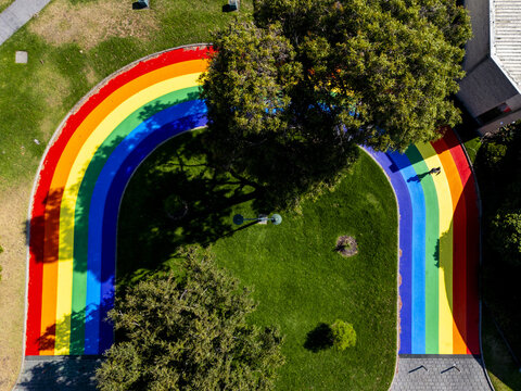 Aerial view of a vibrant rainbow pride pathway curves through lush green lawns, casting playful shadows beneath mature trees, Melbourne, Victoria, Australia.