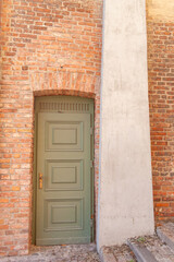 A green door stands at a brick wall, while concrete supports rise beside it in a sunny location. Steps lead up to the entrance.