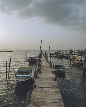 View of weathered wooden pier stretches into calm waters under a cloudy sky, boats bob gently, reflecting the muted light, creating a serene coastal scene, Comporta, Set&uacute;bal, Portugal.