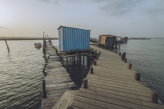 View of weathered wooden pathways stretch over calm waters towards rustic shacks, contrasting the tranquil sea and cloudy sky, Comporta, Set&uacute;bal, Portugal.