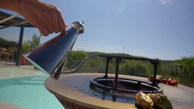A person pours oil from a stainless steel pitcher onto the hot, circular grill cooking plate, which has vegetables (lettuce, tomatoes) already placed on it. Focus on preparation for outdoor cooking.