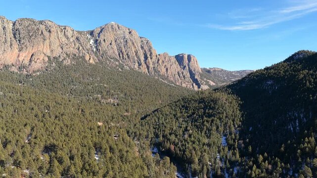 Drone footage advances above a pine-covered mountain valley toward towering rock faces, revealing the Brazos Cliffs rising sharply above dense forest with light snow patches and rugged alpine terrain.