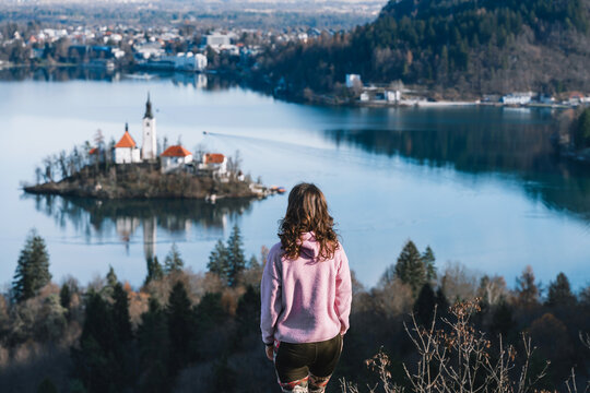 Woman enjoy the scenic view of Bled Lake with Church on Island in middle of lake framed with mountain peaks covered in snow. Lake Bled in winter, Slovenia, Europe. Popular travel destination.