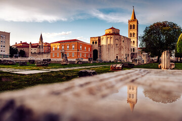 View of ancient ruins and a towering church steeple pierce the skyline, reflected in a still puddle, contrasting old stones with vibrant buildings, Zadar, Zadar County, Croatia.