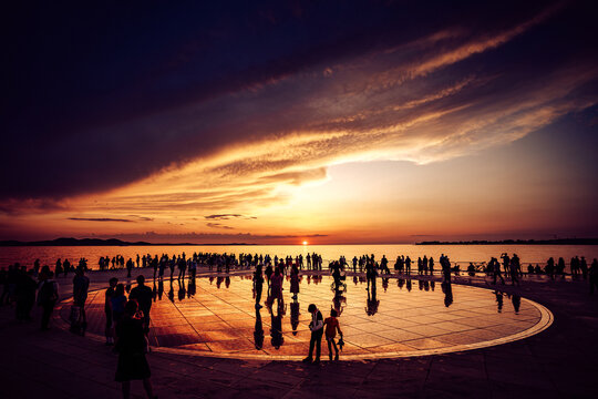 View of a gathering of people silhouetted against a fiery sunset reflected on a large, circular installation by the sea, Zadar, Zadar County, Croatia.