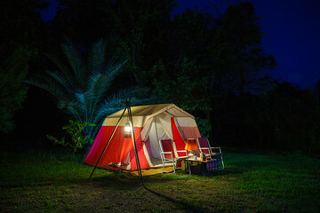 Camping tents at night, vintage cabin tents, antique oil lamps, retro chairs, and a group of camping tents with outdoor coffee-making facilities on wooden tables in a forest camping area.
