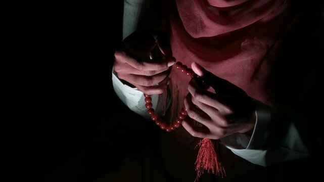 Cinematic close-up of hands holding prayer beads, worn by a young woman in a headscarf, dark studio lighting emphasizing spirituality, belief, reflection, devotion, and emotional depth