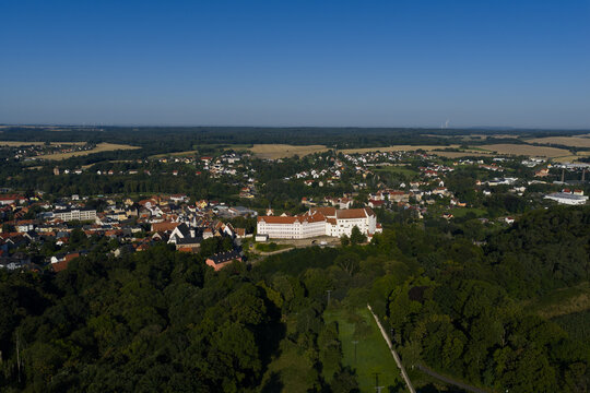Aerial view of Colditz Castle perched atop a wooded hill, overlooking a town nestled amongst verdant forests under a clear blue sky, Colditz, Sachsen, Germany.