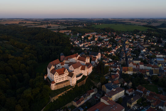 Aerial view of Colditz Castle's aged stone walls contrasting with the lush green forest beneath a tranquil sky, Colditz, Sachsen, Germany.