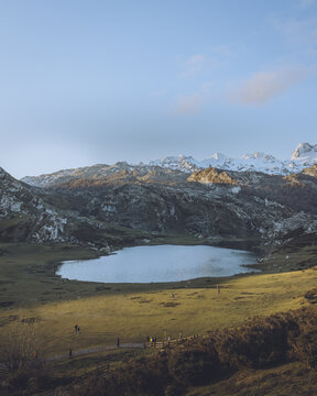 View of the tranquil lake mirroring the snow-capped peaks under a serene sky, embraced by the vibrant green meadow, Lagos de Covadonga, Cangas de Onis, Asturias, Spain.