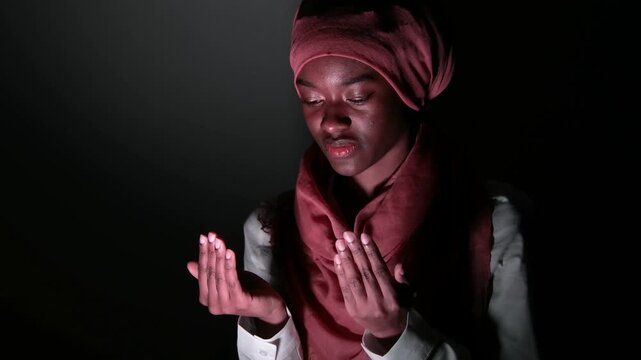 Cinematic low-key portrait of a young woman in a headscarf raising her hands in prayer, dark studio atmosphere highlighting spirituality, belief, emotional depth, reflection, and quiet devotion