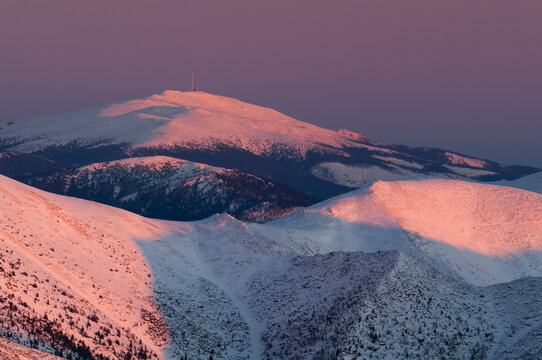 Aerial view of snow-covered peaks kissed by the soft blush of dawn, with a distant structure atop the highest point, Nizke Tatry, Banska Bystrica Region, Slovakia.