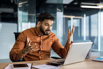 Young businessman actively participating in a video call, talking and gesturing with hands while using a laptop at his office desk during an online meeting or presentation