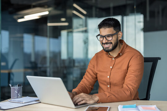 Young smiling indian businessman engaging with his laptop while happily working in a contemporary corporate office environment, embodying professionalism, success, and digital productivity