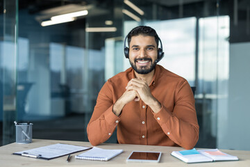 Smiling indian man wearing a headset and orange shirt sitting at his desk in a modern office, engaged in remote work, online meeting, or providing customer support