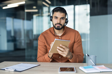 Young bearded man in headset with microphone on a video call, working from home office, focused and taking notes in a notebook at his desk during a virtual meeting