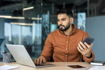 Young indian businessman working on his laptop and holding a smartphone, actively managing multiple tasks and communicating in a modern office environment