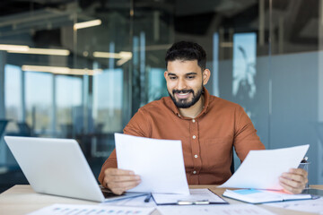 Obraz premium Young businessman analyzing financial documents and reports at his office desk, confidently managing paperwork and working efficiently with a laptop in a corporate environment