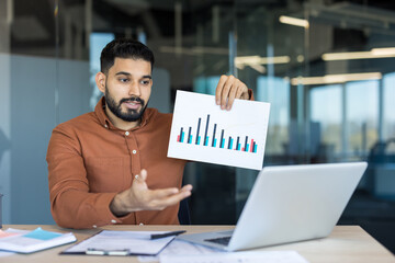 Man holding and presenting a bar chart to colleagues during a remote video conference, discussing business data and financial statistics in a modern office setup