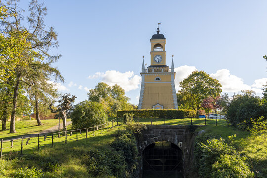 View of Amiralitetsklockstapeln clock tower standing tall above a moss-covered stone tunnel, framed by vibrant green foliage under blue sky, Karlskrona, Blekinge County, Sweden.