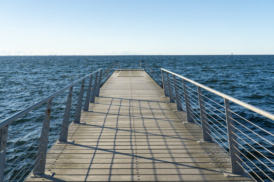 View of a wooden pier stretches out into the shimmering blue sea under a clear sky, casting long shadows, Vastra Hamnen, Malmo, Sweden.