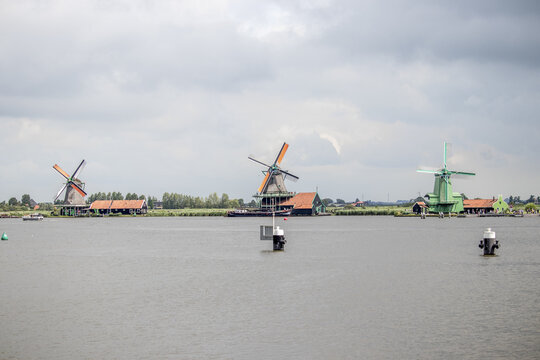 View of windmills standing steadfast against the water, their colorful tops contrasting with the serene water and overcast sky, Zaanse Schans, Noord-Holland, Netherlands.