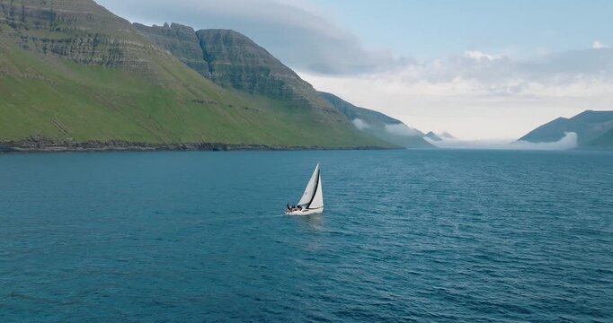 Aerial view of a lone sailboat cutting through the deep blue water, set against the backdrop of towering green mountains, Klaskvik, Faroes.