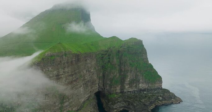 Aerial view of majestic cliffs dramatically meeting the ocean, lush green grass contrasting with the rugged rock formations, shrouded in mist, Kalsoy, Faroes.
