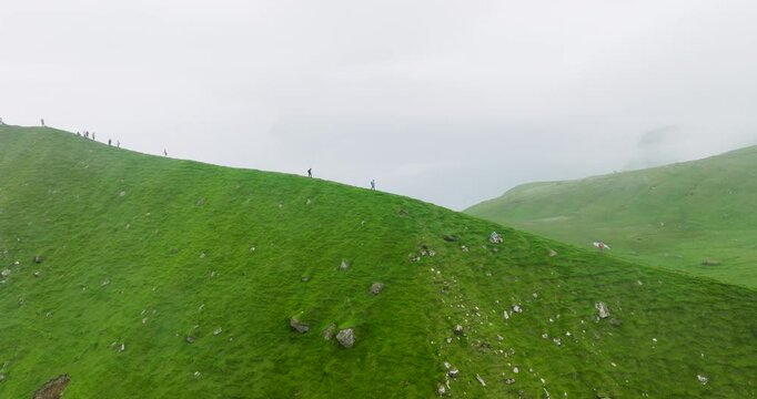 Aerial view of vivid green hills contrasting with the grey sky, with tiny figures of people walking along the ridge, Kalsoy, Faroes.