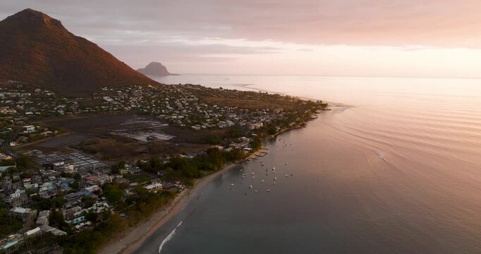 Aerial view of a coastal town nestled against a mountain, with boats dotting the water under a pastel sunset, Riviere du Rempart, Mauritius.