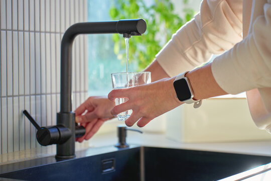 Hand filling glass with drinking water from kitchen faucet in modern home interior. Woman pouring clear water from tap filter. Concept of drinking water, daily hydration and quenching thirst