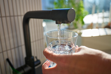 Hand filling glass with drinking water from kitchen faucet in modern home interior. Woman pouring...