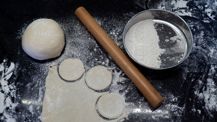 Rolled dough circles, rolling pin, flour sieve and fresh dough ball on dark kitchen background. Traditional cooking concept, rustic food preparation, homemade cuisine.
