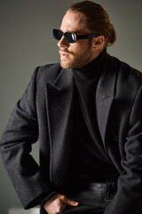 Obraz premium Handsome man in stylish sunglasses and elegant coat poses confidently in grey studio